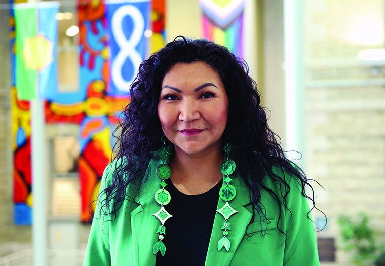 Margaret Hart in the Brodie Atrium at Bannatyne campus, behind her are flags and an Indigenous mural.