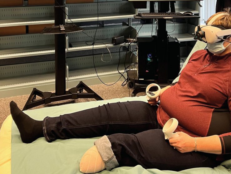 A patient partner sits up on a bed and is wearing a VR headset and is holding two VR controllers.